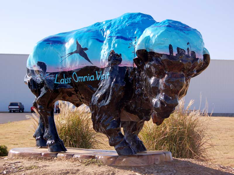 Painted buffalo sculpture with prairie horizon and sunrise. Downtown Bartlesville skyline. Local animal silhouettes dot horizon.