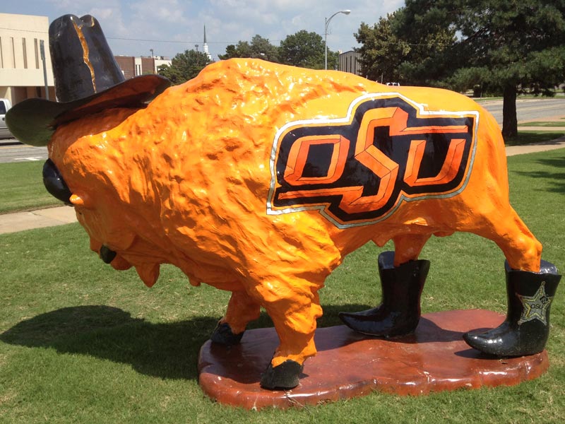 Orange painted buffalo sculpture wearing a black cowboy hat, Oklahoma State University logo on body and black cowboy boots on back legs.