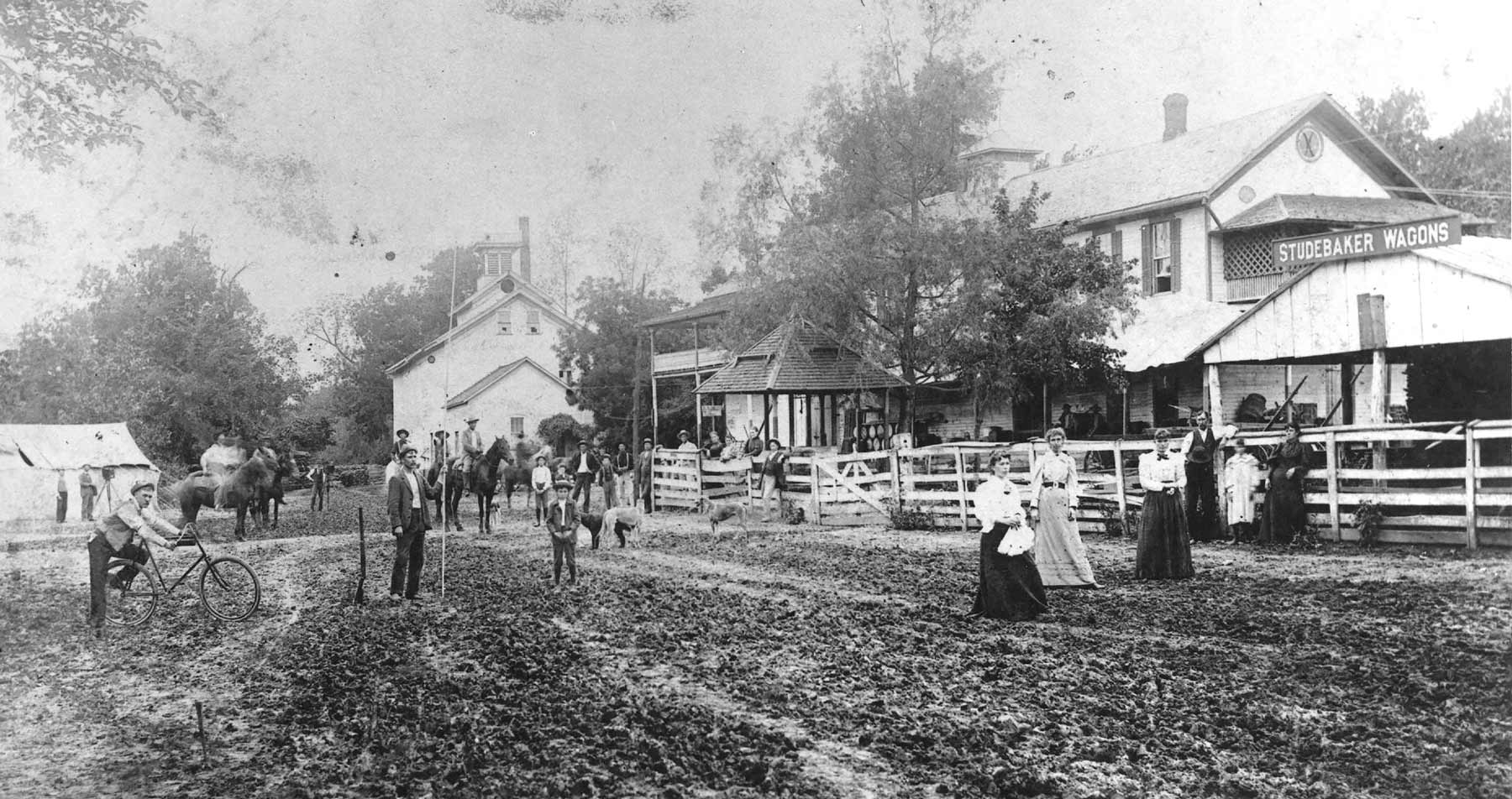 Crowd gathered on muddy dirt road in front of Bartles store/home with millsite in background